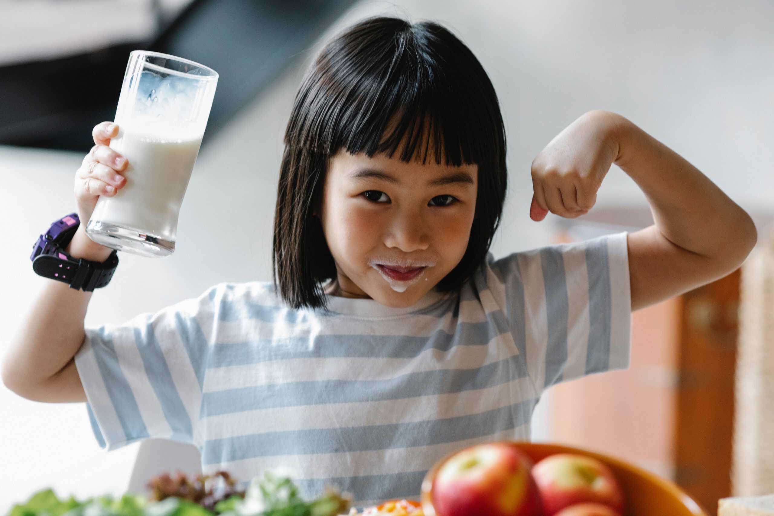 Mała dziewczynka siedząca przy stole ze szklanka mleka w ręce i wąsami od napoju wokół ust